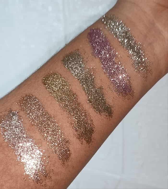 Six glitter eyeshadow swatches applied in a row on a person's forearm, showing shimmering shades of silver, bronze, gold, and lavender-purple.